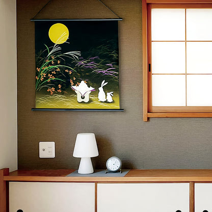 Decorative wall hanging with two white rabbits under a yellow moon on a dark background, placed above a wooden desk with a lamp and clock.
