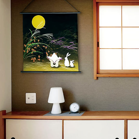 Decorative wall hanging with two white rabbits under a yellow moon on a dark background, placed above a wooden desk with a lamp and clock.